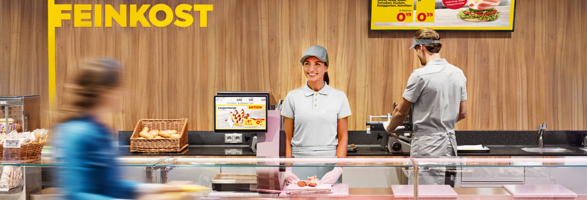 Eine lächelnde Mitarbeiterin steht an der Feinkosttheke.