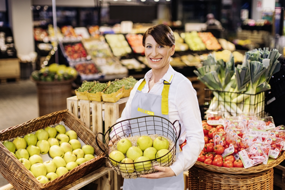 Eine lächelnde Mitarbeiterin steht mit einem Obstkorb in der Frischeabteilung.
