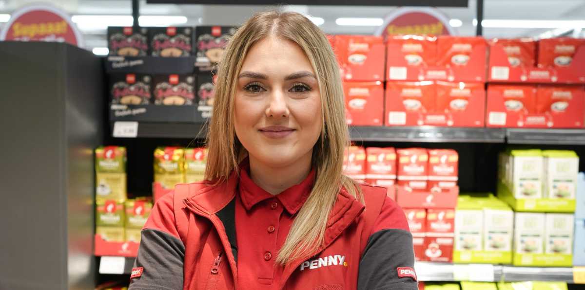 Eine lächelnde Mitarbeiterin steht vor einem Regal.